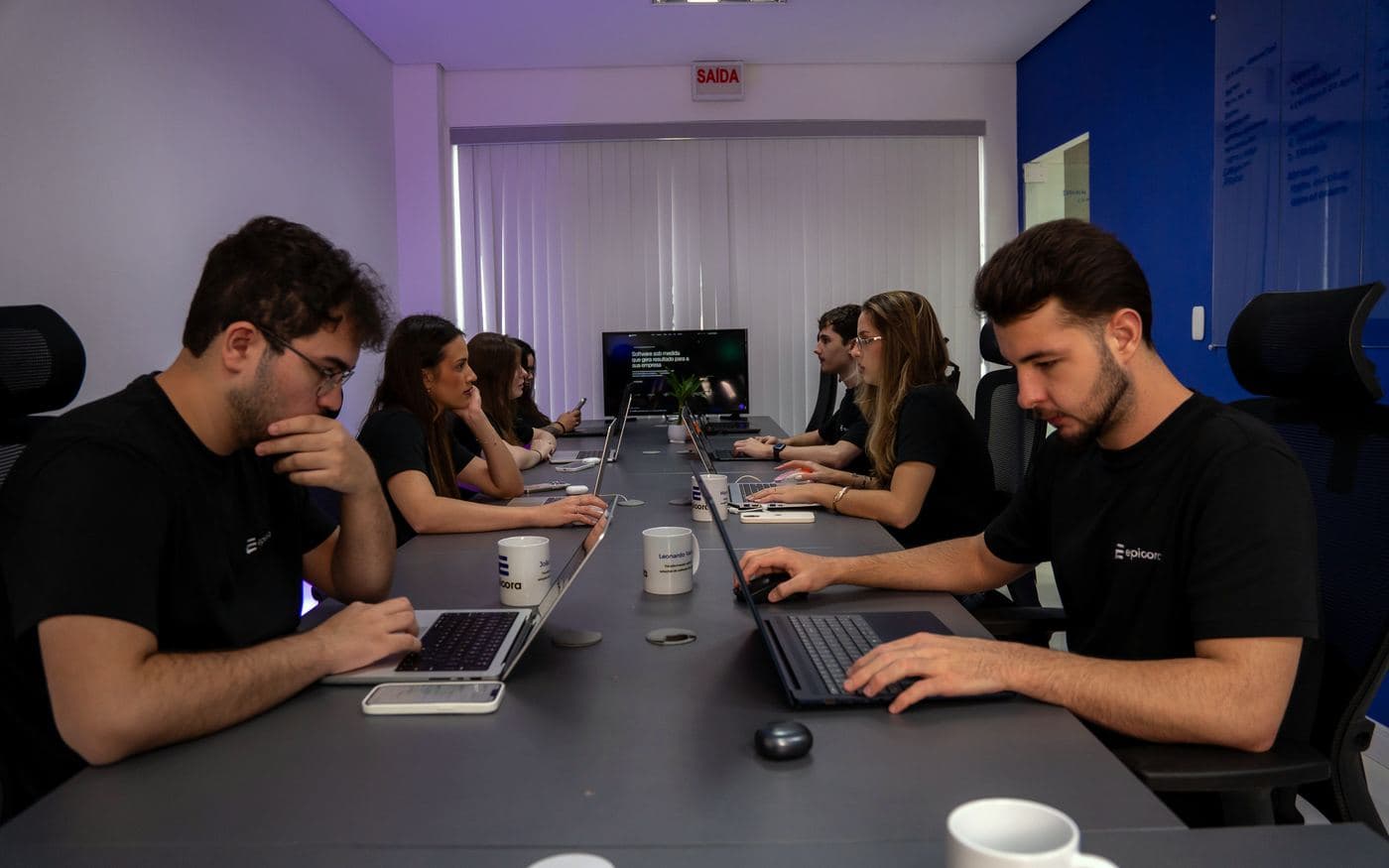 Escritório da Epicora: integrantes da equipe trabalham concentrados nos notebooks ao longo da mesa comprida, com a parede azul à direita.